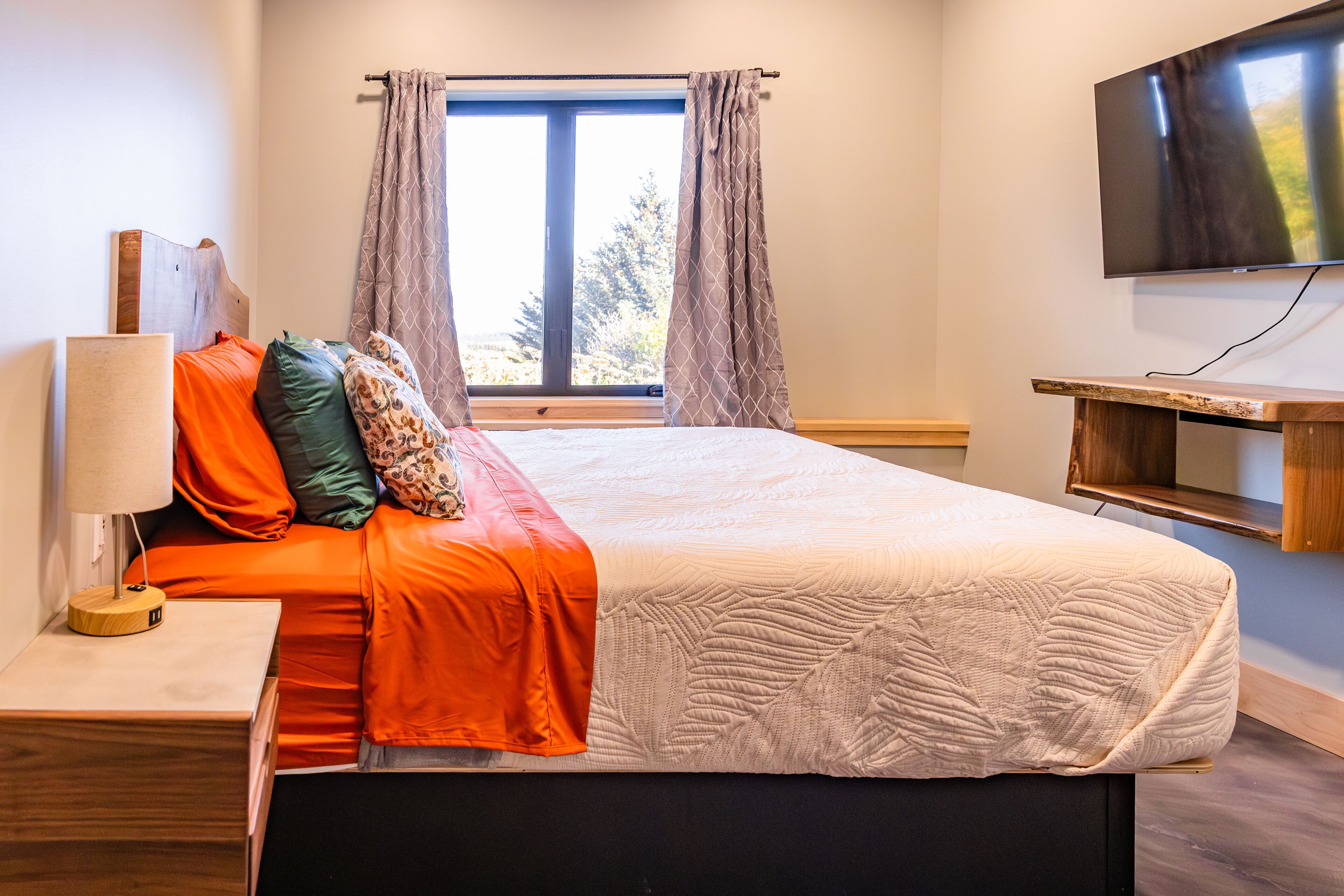 Une chambre à coucher avec un lit, une table de chevet, une lampe, un téléviseur et une fenêtre avec des rideaux.