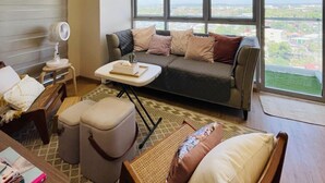 Una sala de estar con sofá, reposapiés y una mesita de centro, un ventilador y una ventana con vistas a la ciudad.
