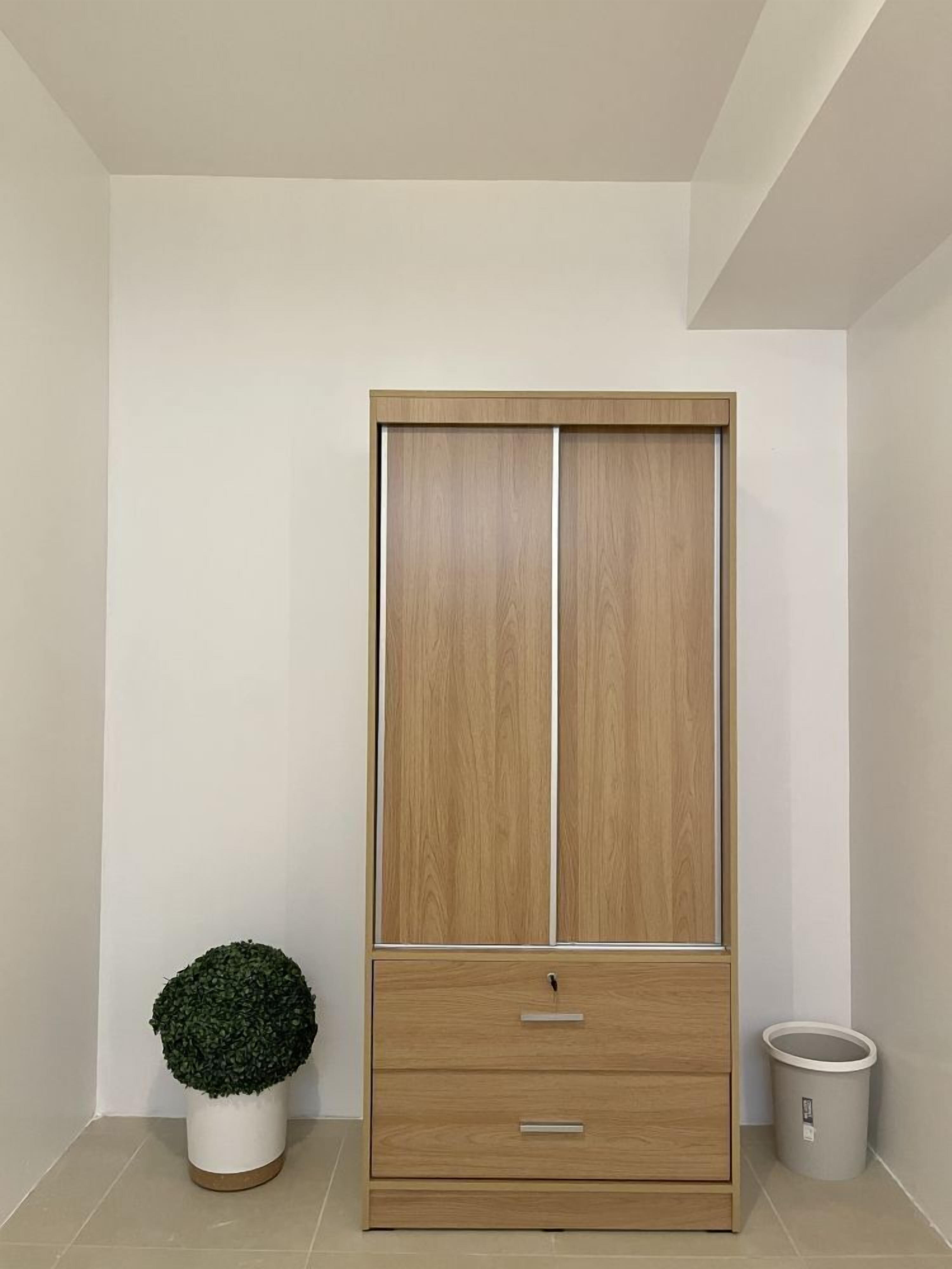 Une grande armoire en bois, dotée de deux tiroirs et d’une grande porte, accompagnée d’une plante en pot et d’une poubelle.