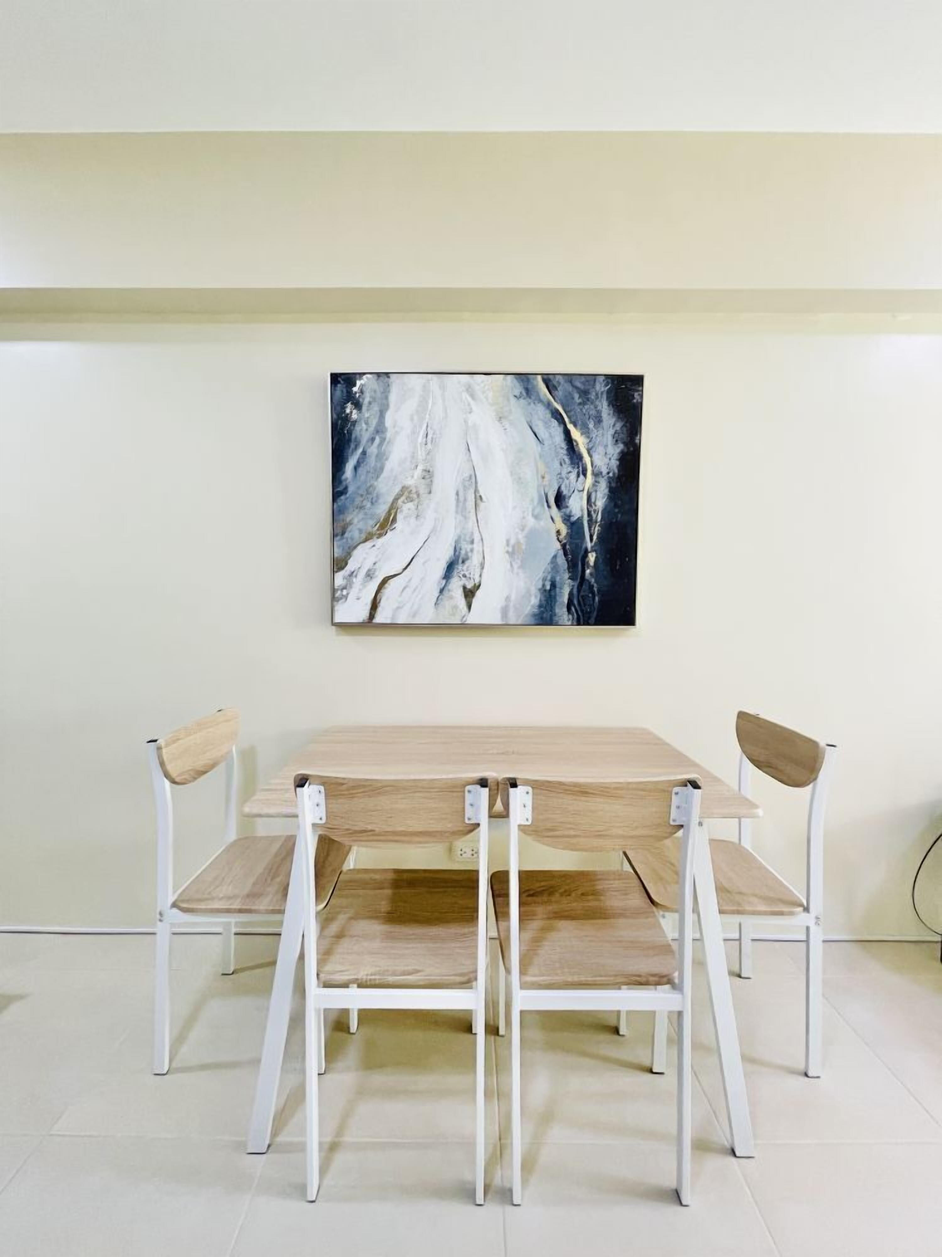 A dining area with a wooden table and white chairs, a large abstract painting on the wall, and a light-colored floor.