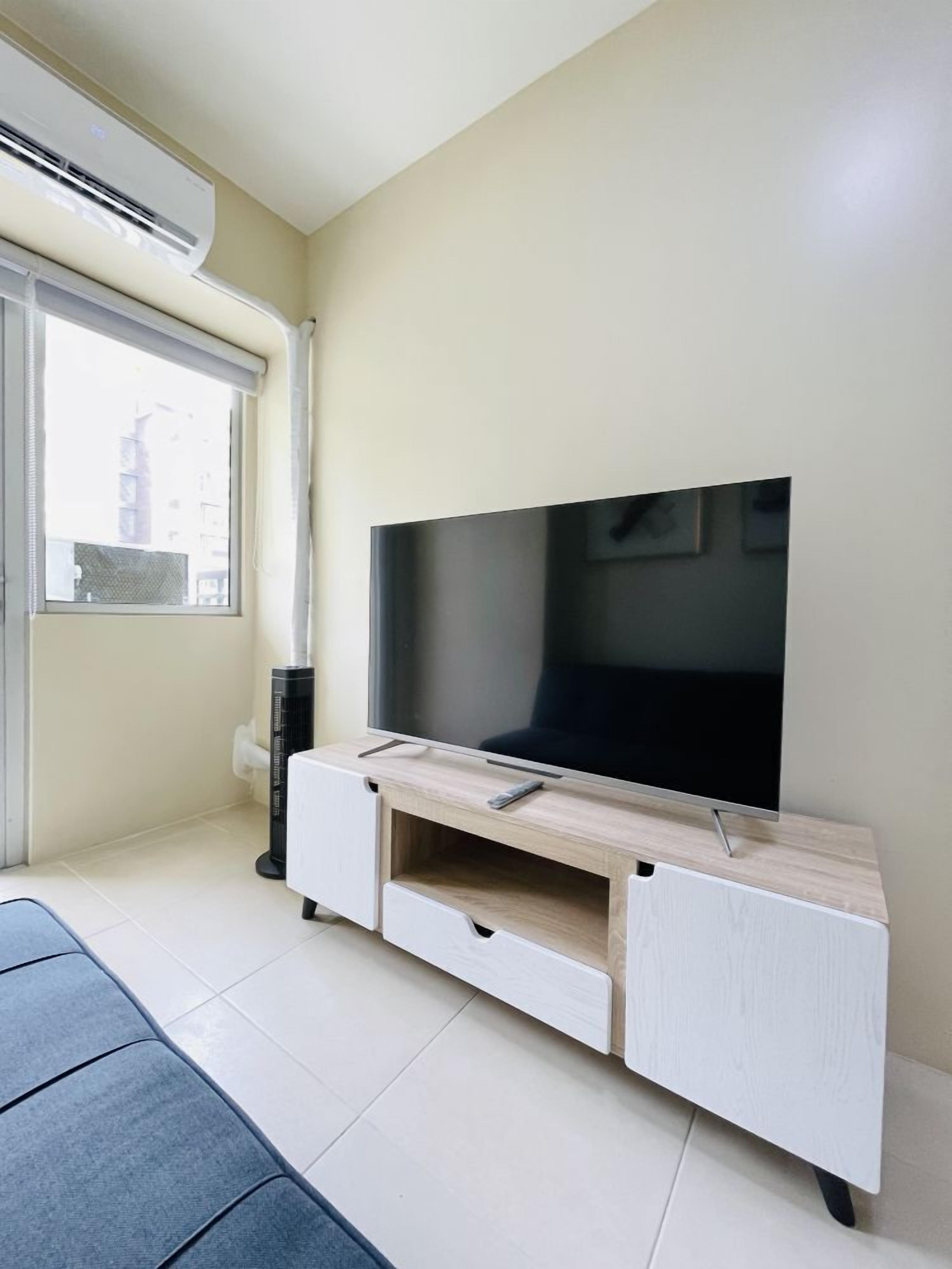 A modern living room with a flat-screen TV on a wooden TV stand, a small heater, and a window with blinds.