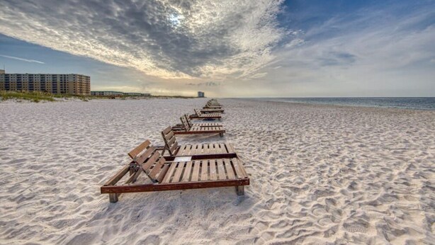 A beach with rows of wooden lounge chairs facing the ocean, a clear sky, and a distant building. - Plantation West 1367 (Gulf Shores)