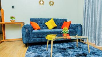 Una sala de estar con un sofá azul, cojines naranja y amarillos, una mesa de centro de vidrio y una alfombra mullida azul.