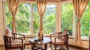 A room with a large window, two wooden chairs, a small table with a bottle and flowers, and a patterned cushion. - Jungle Ghar  (Pipariya)