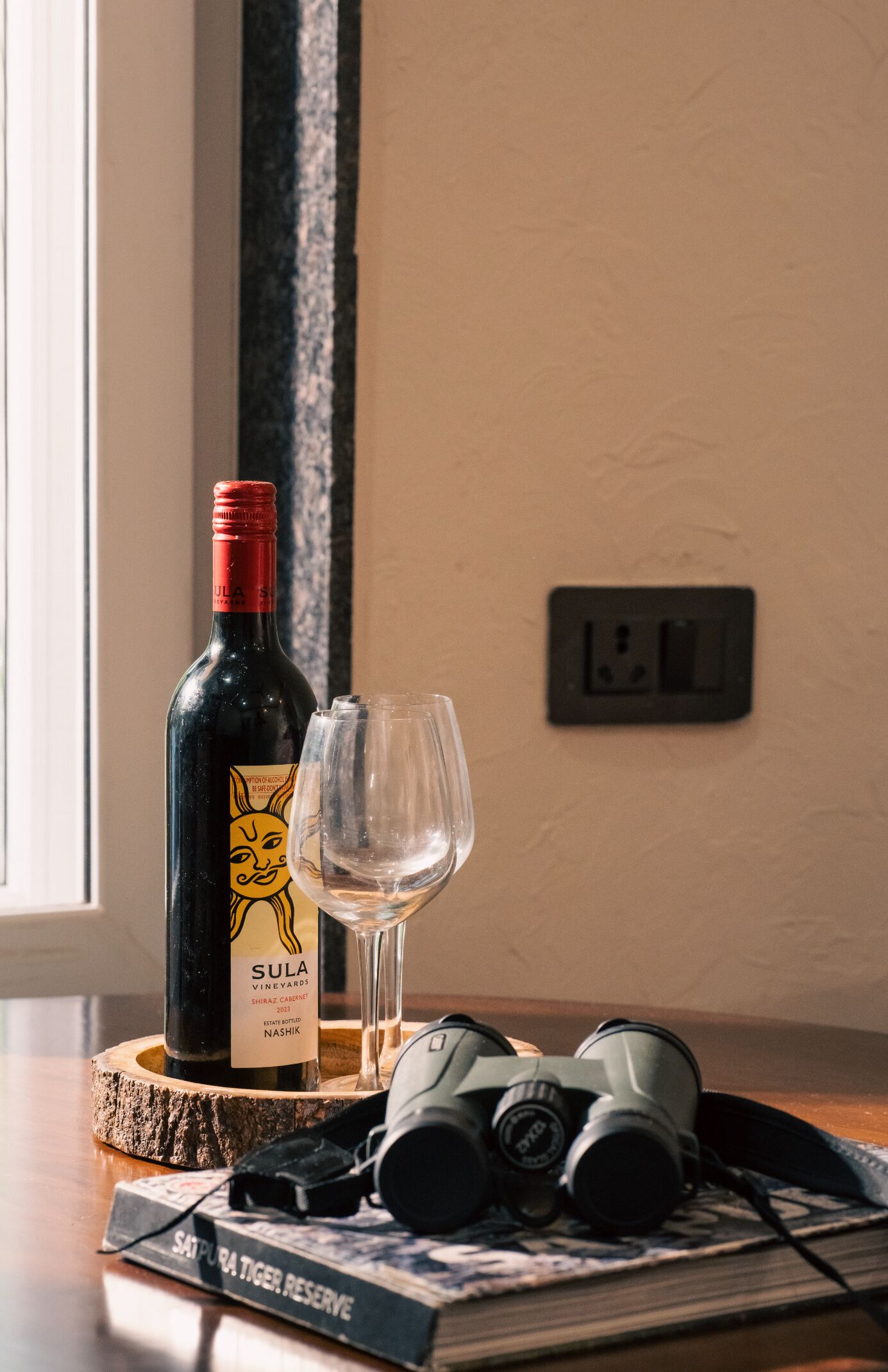 Una botella de vino Shiraz Cabernet Sauvignon 2023 de Sula Vineyards, una copa de vino, unos binoculares y un libro titulado 'Satpura Tiger Reserve' sobre un posavasos de madera.