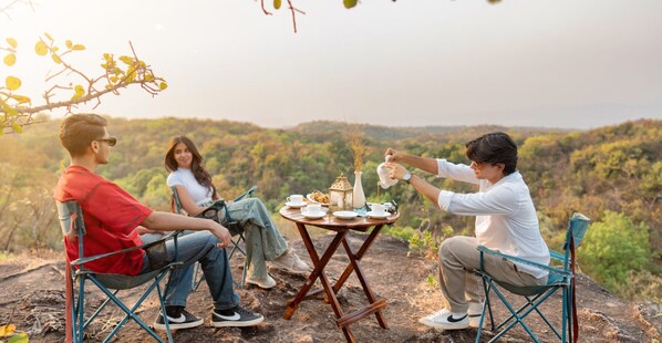 Trois personnes sont assises à une table en bois à l’extérieur, avec un paysage pittoresque en arrière-plan.