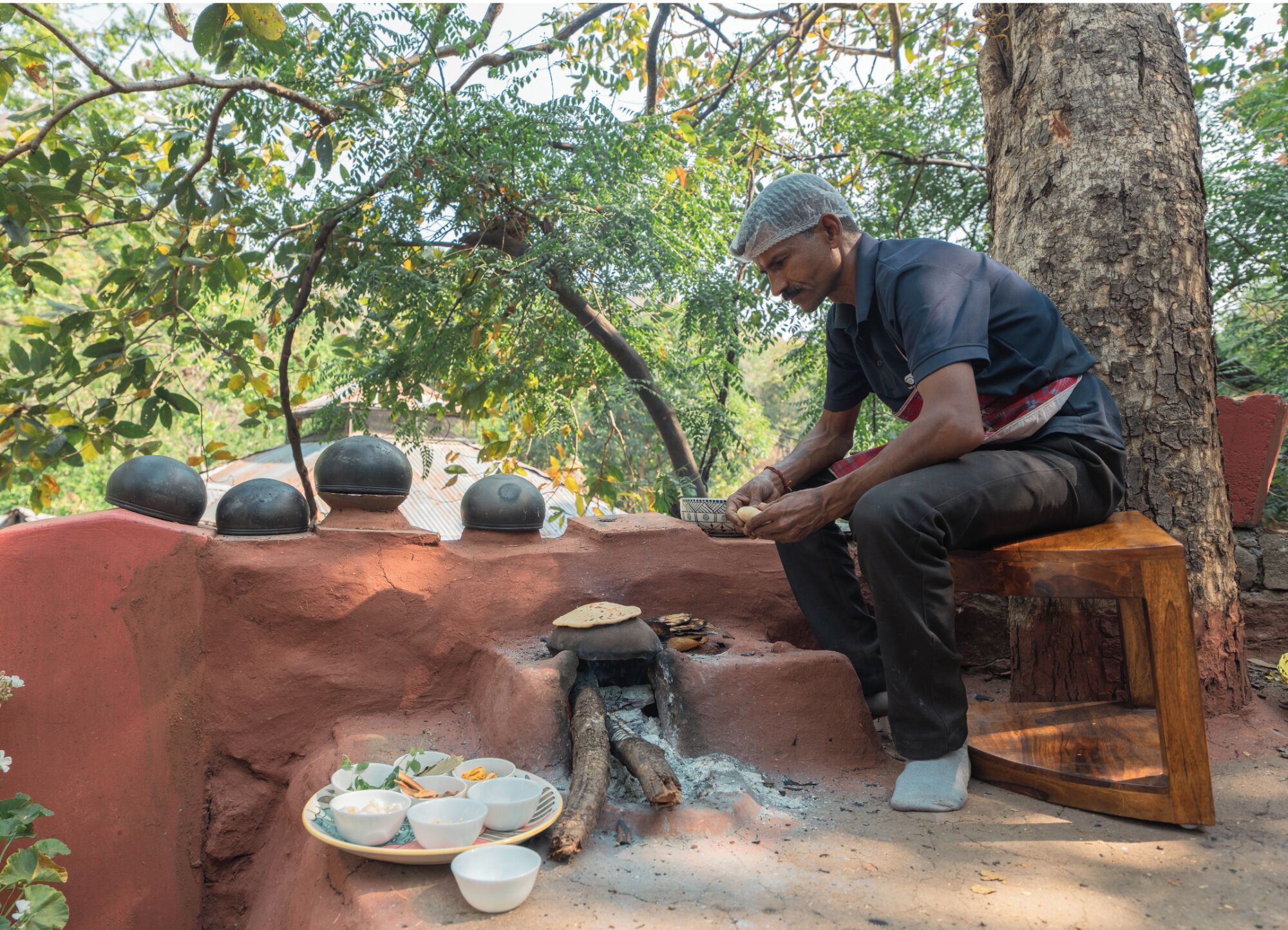 A person sitting by a traditional cooking setup outdoors, surrounded by trees and a red wall.