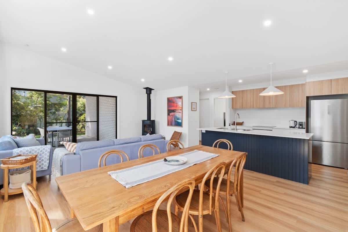 Una cocina moderna con una isla central, una zona de comedor con mesa y sillas de madera, y una sala de estar con sofá y chimenea.