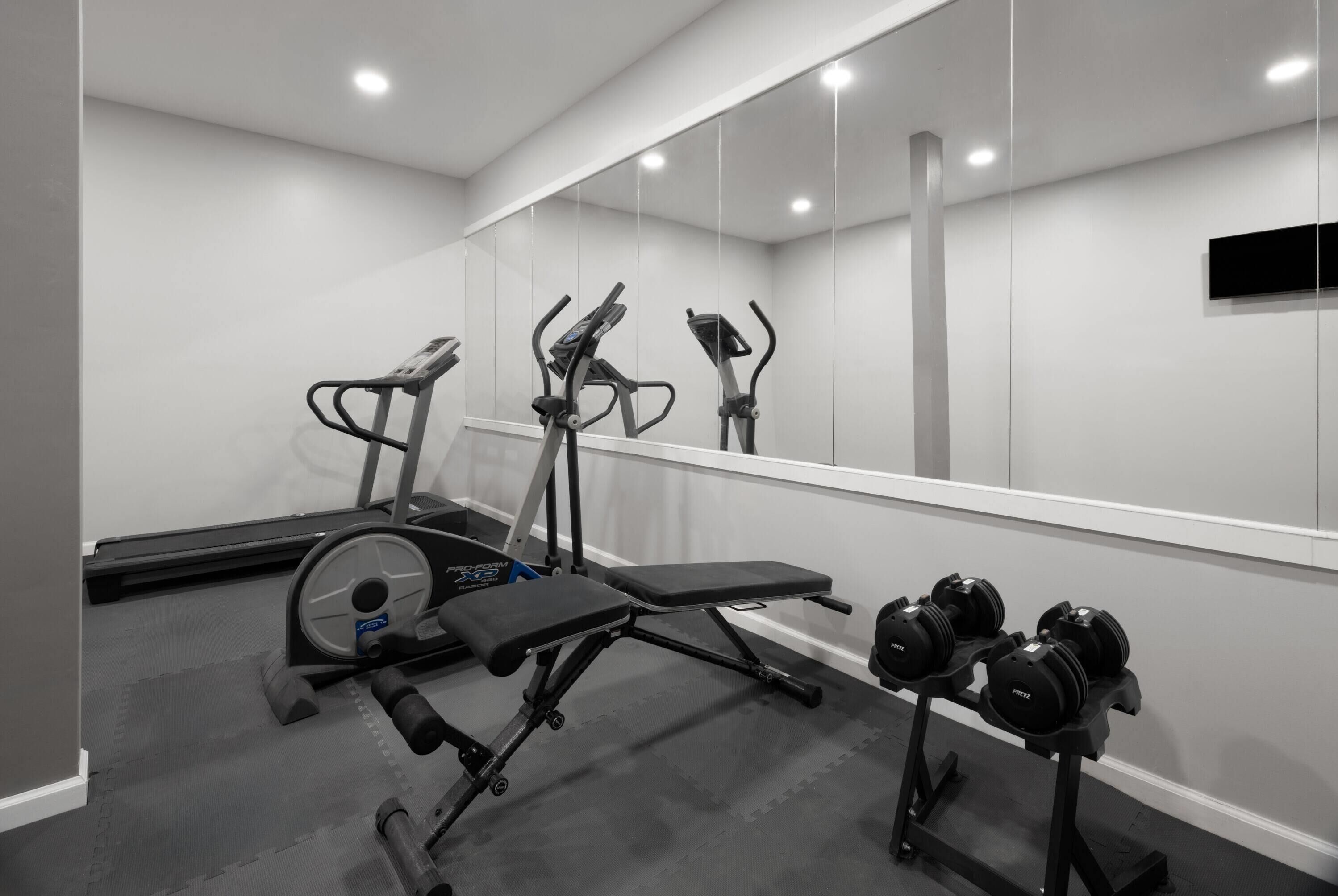 Une salle de sport équipée de matériel d’exercice, notamment un tapis de course, un vélo elliptique, un banc et des haltères.