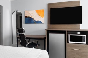 A hotel room with a bed, a desk, a chair, a flat-screen TV, a microwave, a refrigerator, and a painting on the wall.