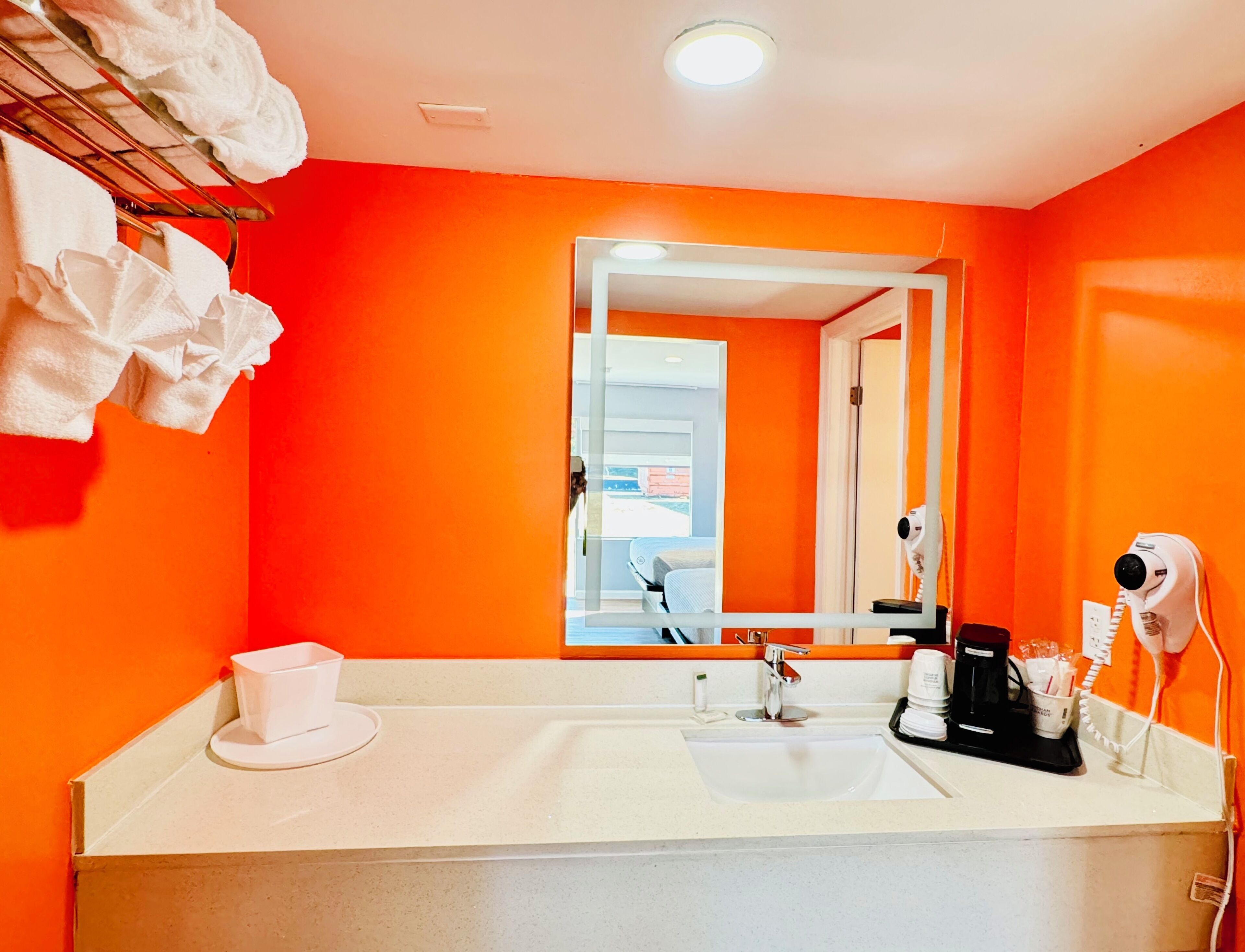 Ein Badezimmer mit orangefarbener Wand, einem weißen Waschbecken, einem Spiegel, einem Handtuchhalter, einem Fön und einer Tasse.