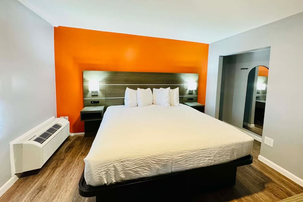 A hotel room with a large bed, an orange accent wall, a wooden floor, a white air conditioner, and a nightstand with a lamp.