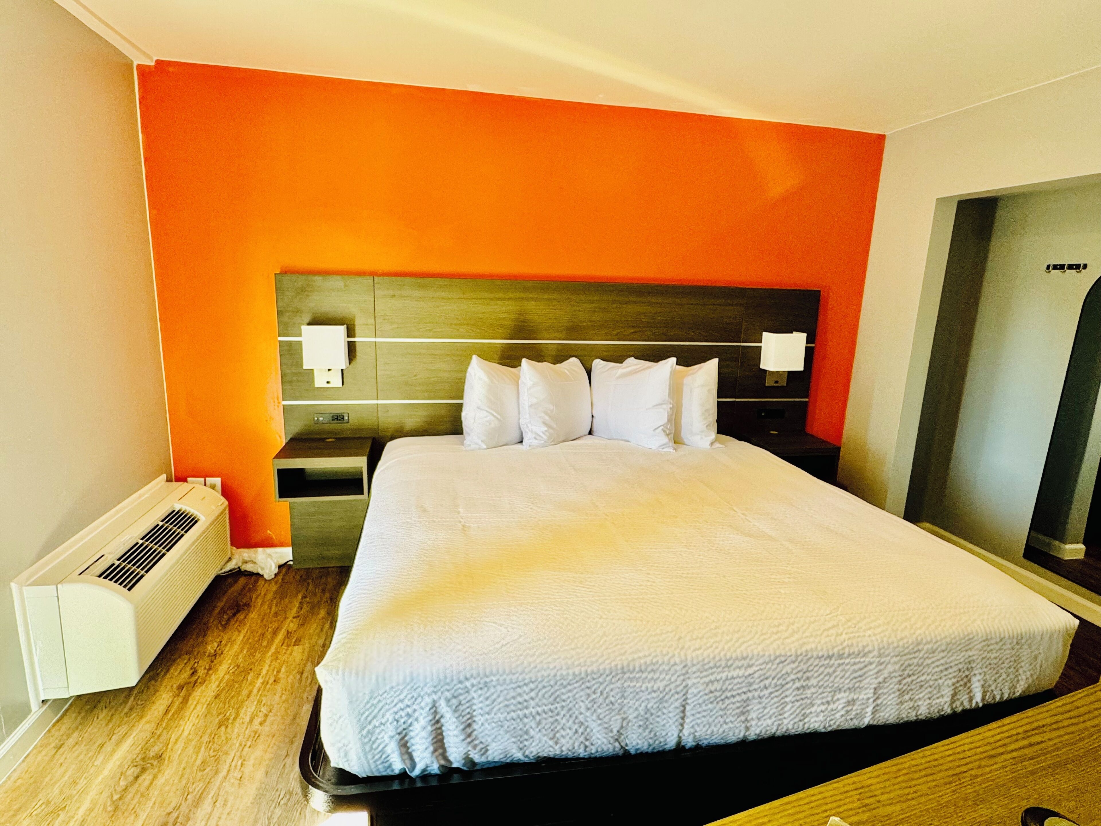 A hotel room with a large bed, a wooden headboard, a nightstand, a wall-mounted lamp, and a heater.