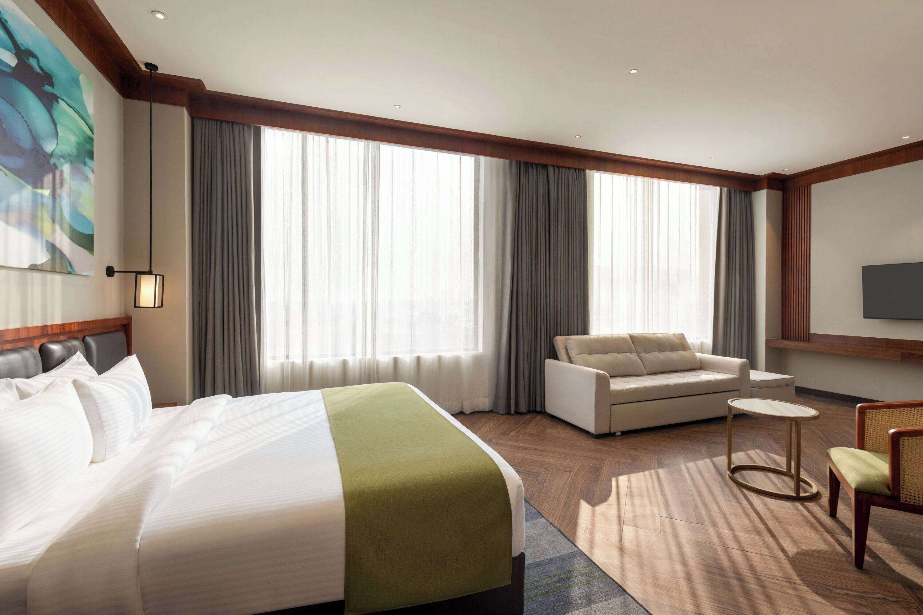 A hotel room with a large bed, a sofa, a small round table, and a TV mounted on the wall.
