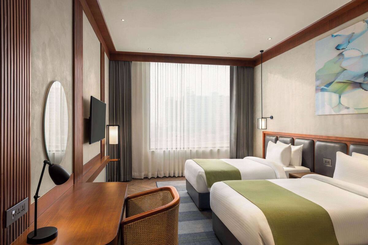 A hotel room with two beds, a desk with a chair, a TV, and a large window with curtains.