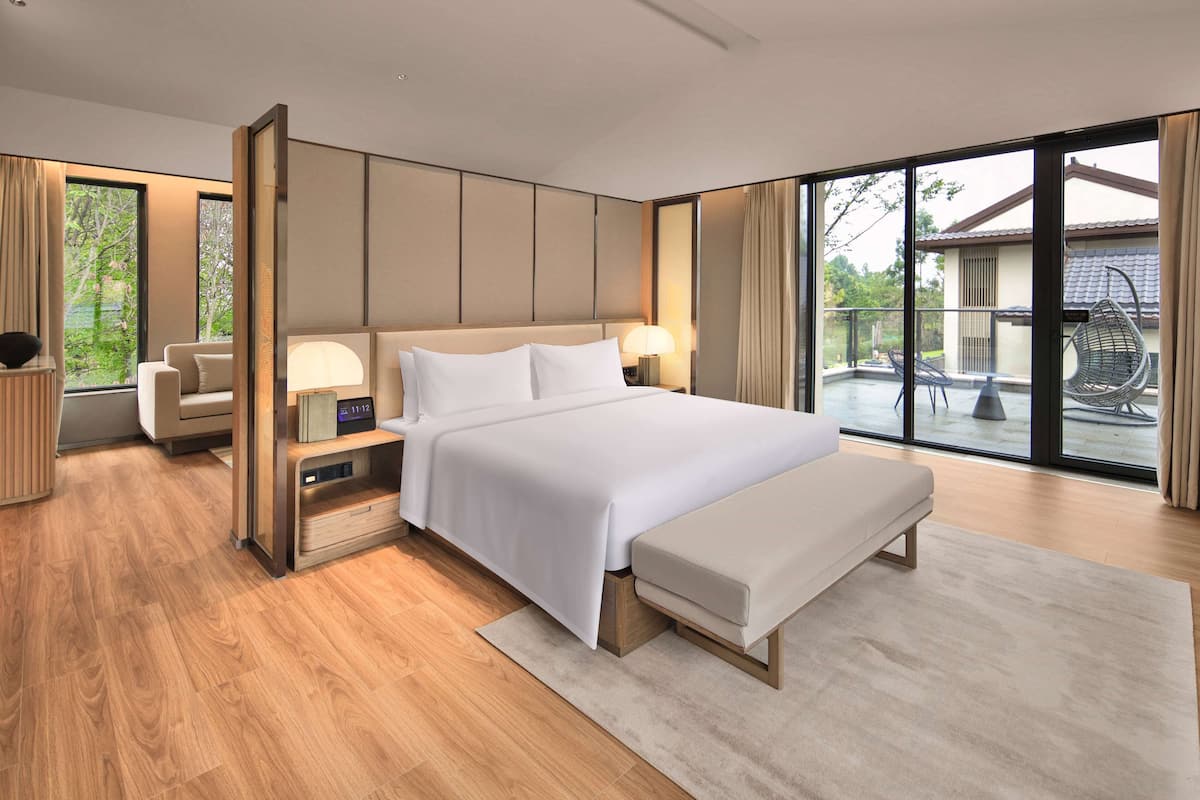 Une chambre d’hôtel moderne, dotée d’un grand lit, de tables de chevet, d’un banc et offrant une vue sur l’espace extérieur.