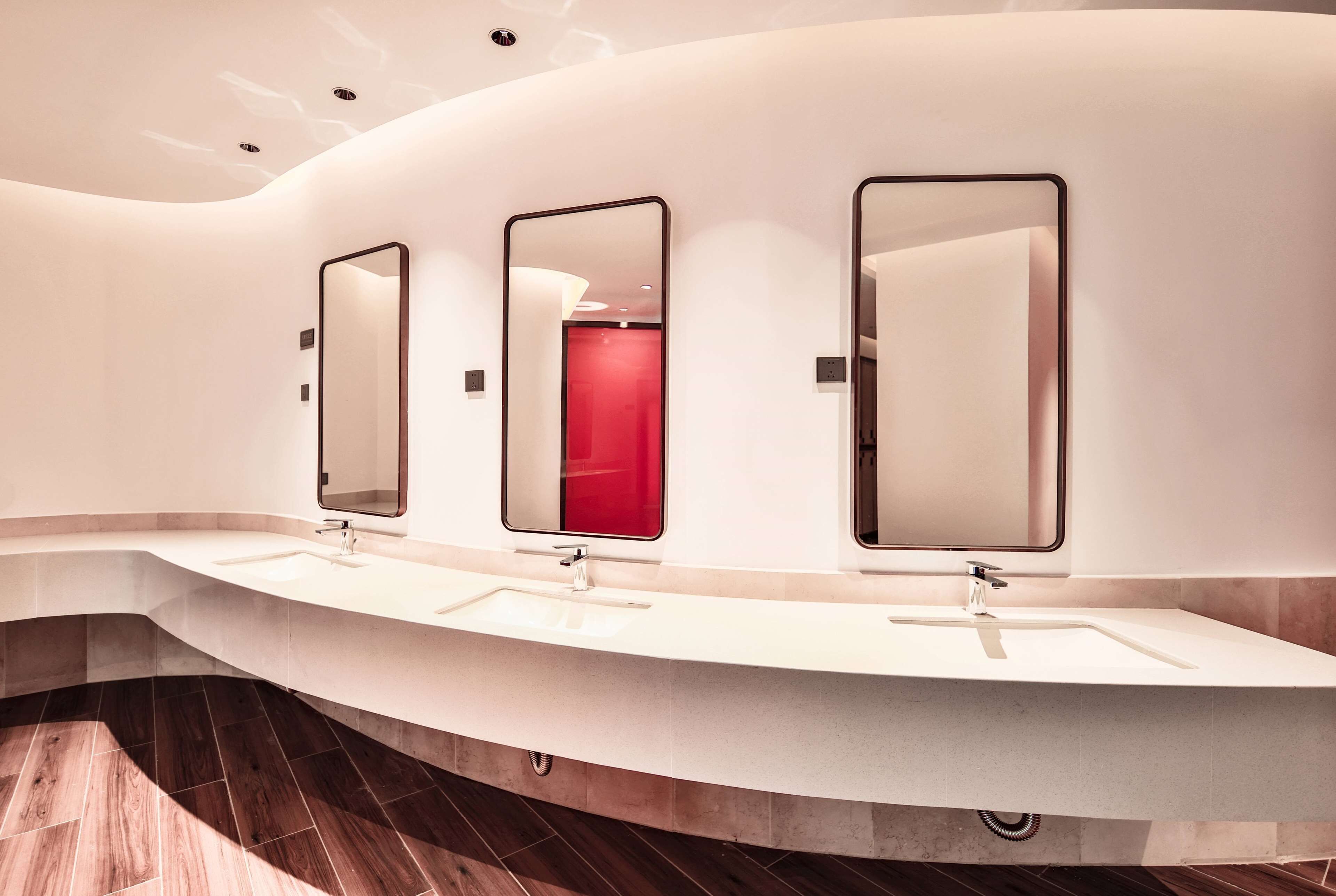 A modern bathroom with three sinks and mirrors, a curved design, and a red door in the background.