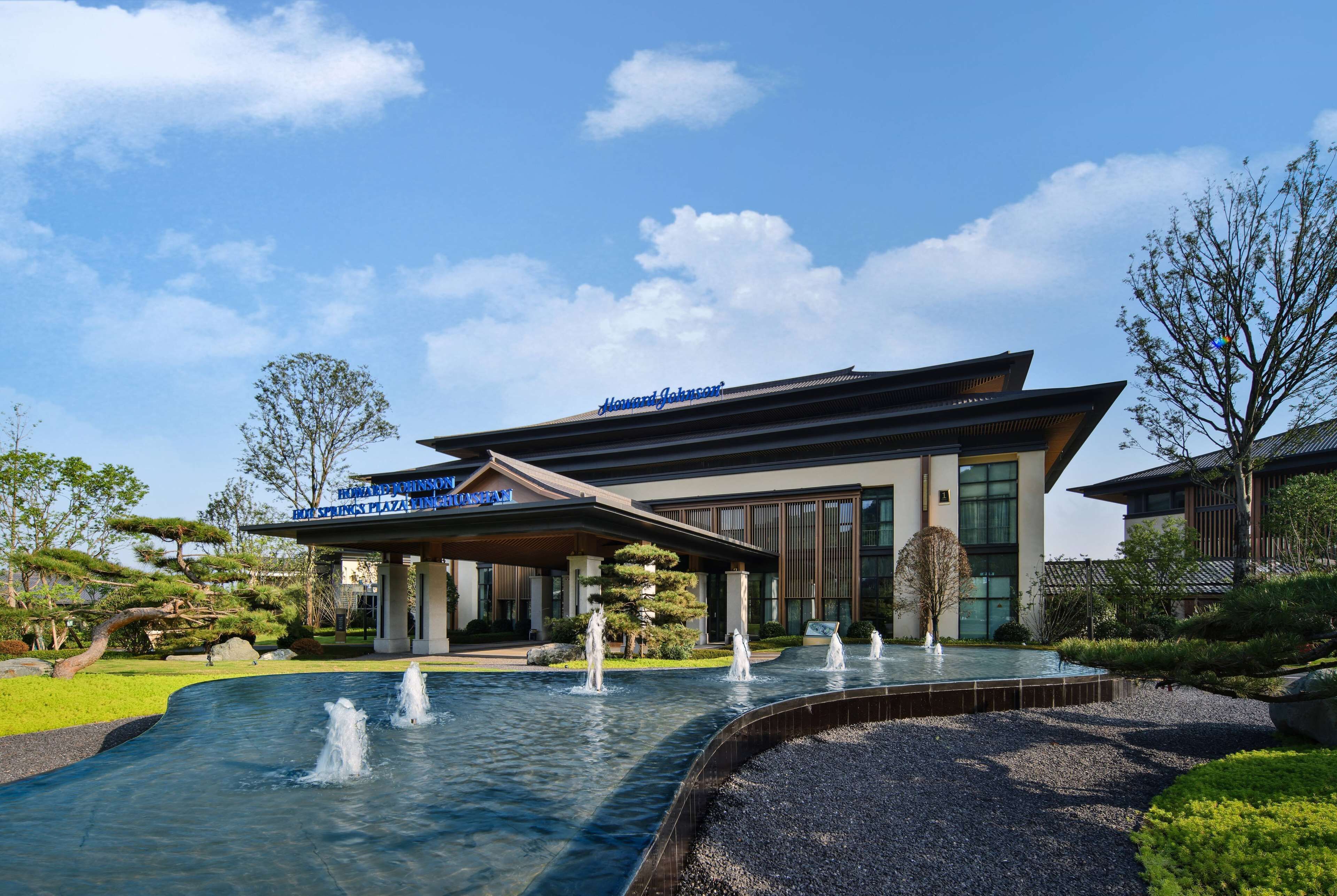 A modern hotel with a water feature in front, a covered entrance, and a traditional-style building in the background.