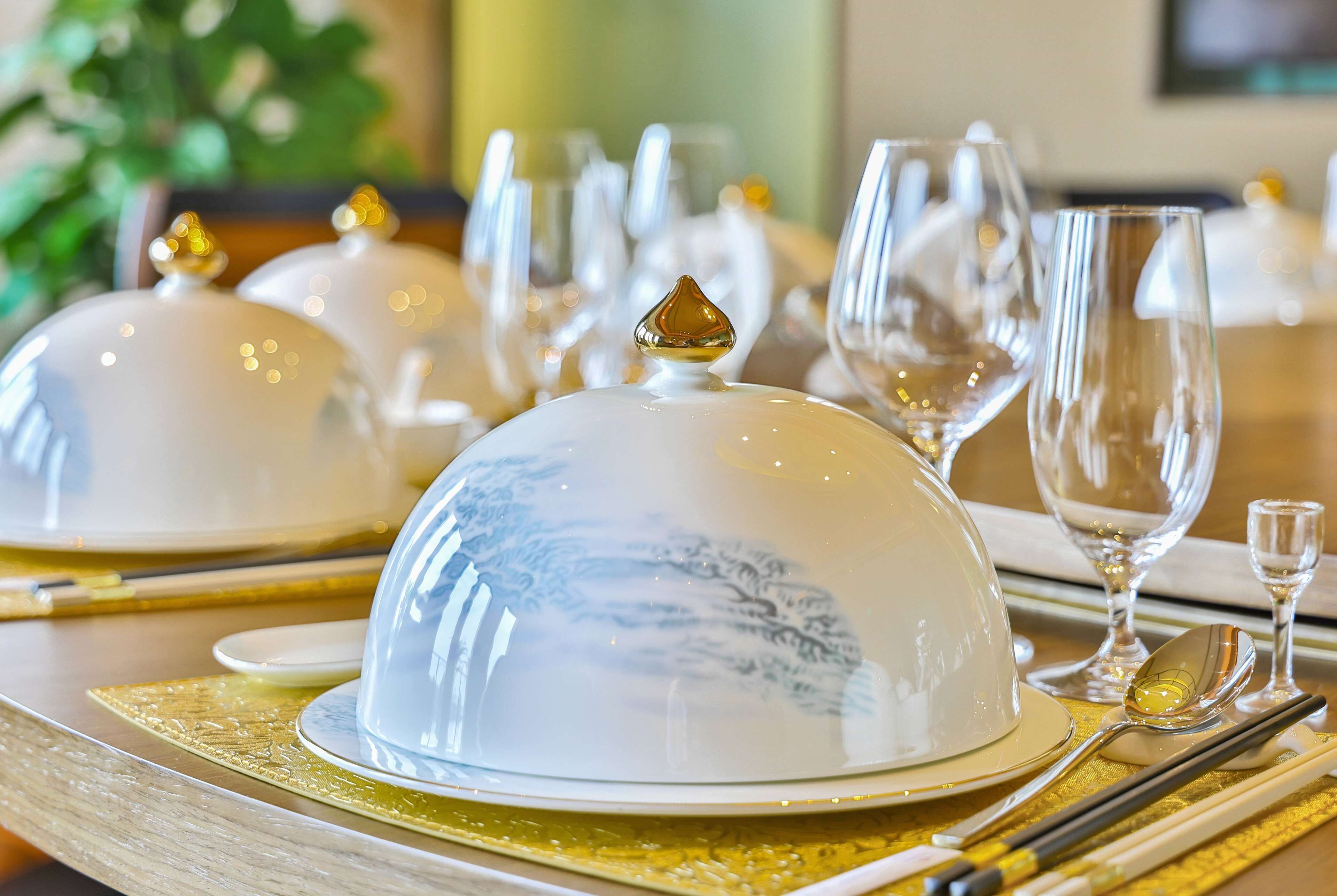 A table set with white dinnerware, including plates, bowls, and a covered tureen, with a gold placemat underneath.