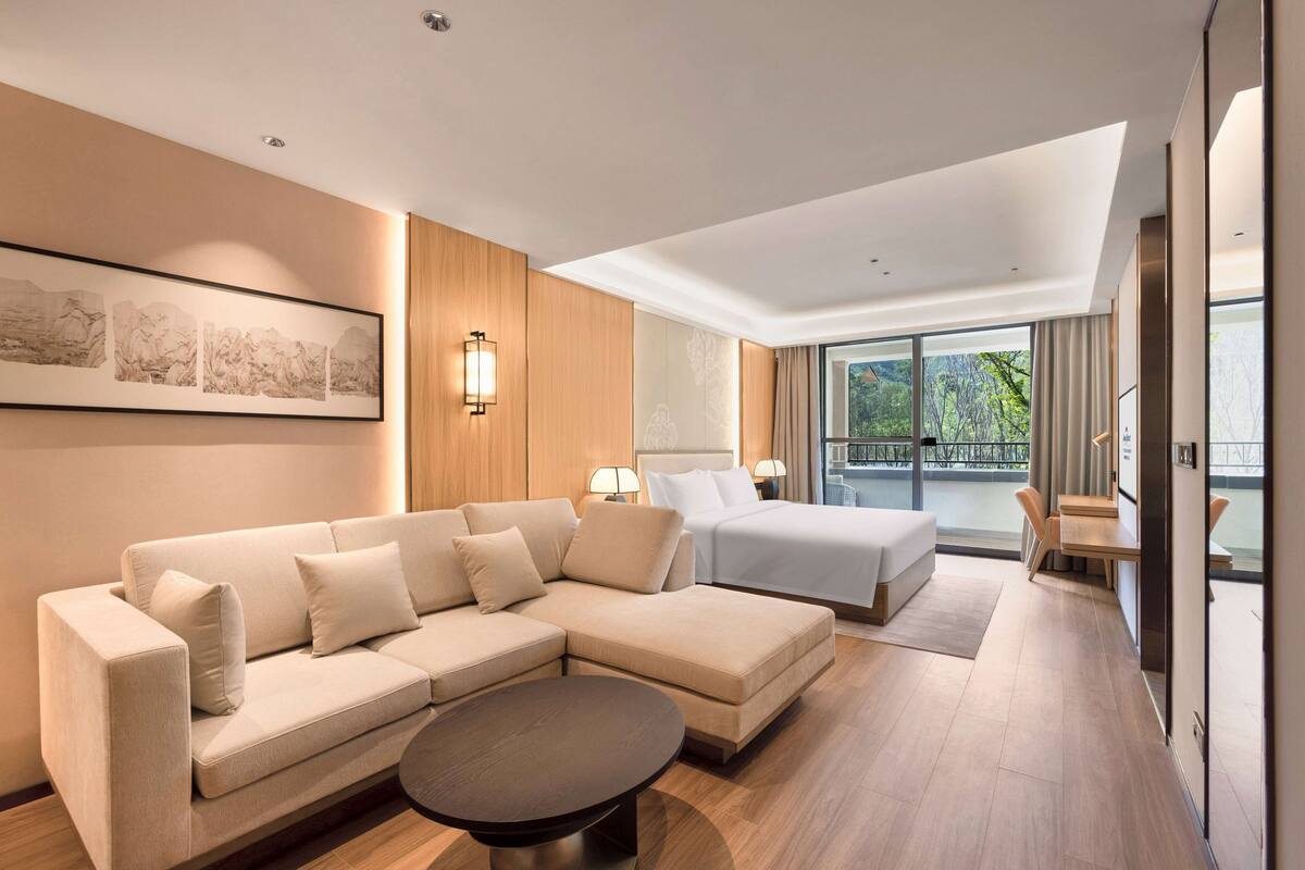 Une chambre d’hôtel moderne dotée d’un canapé d’angle, d’un lit, d’une petite table et d’une grande fenêtre donnant sur l’extérieur.