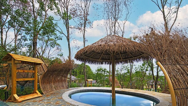A circular stone pool with a thatched umbrella, surrounded by stone tiles and wooden structures.