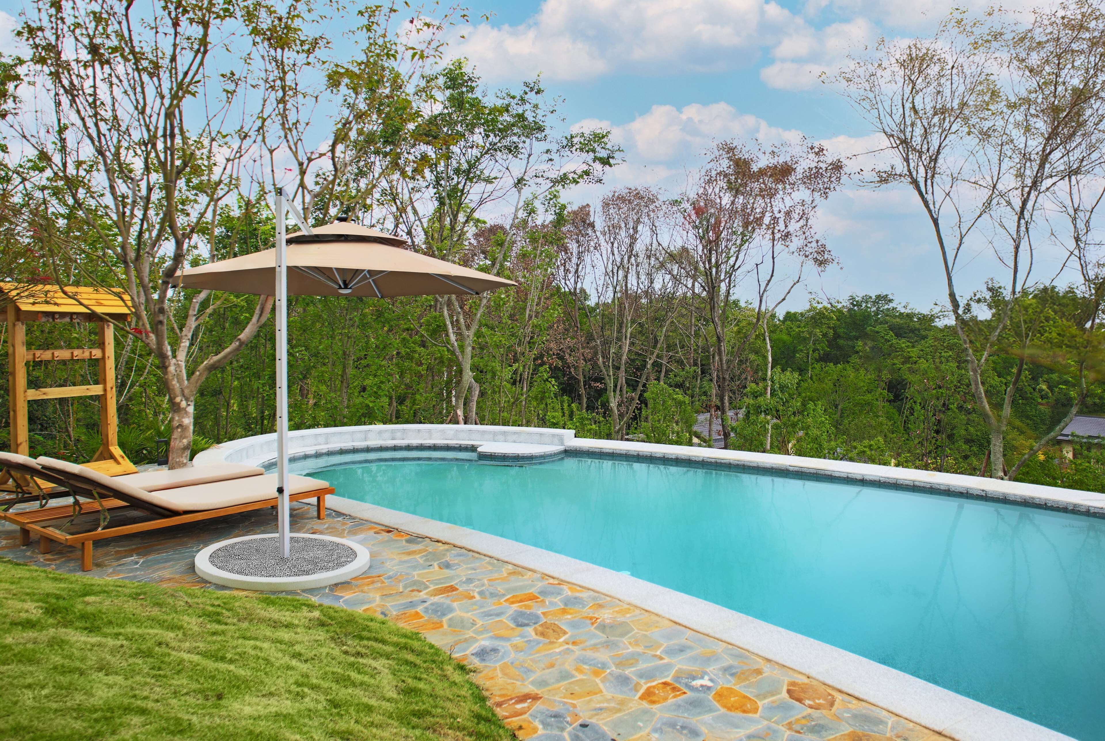A swimming pool with a stone deck, surrounded by lush greenery and a wooden deck with lounge chairs.