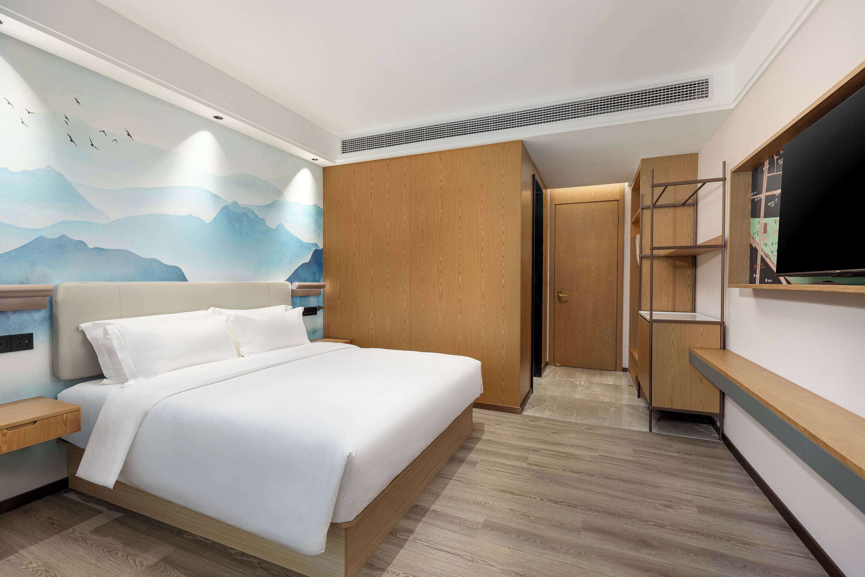 Une chambre d’hôtel moderne dotée d’un grand lit, d’une armoire en bois, d’un téléviseur intégré et d’une fresque représentant des montagnes.