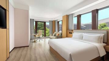 Ein modernes Hotelzimmer mit einem großen Bett, einem Schreibtisch und einem Sessel. Große Fenster bieten einen Blick ins Freie.