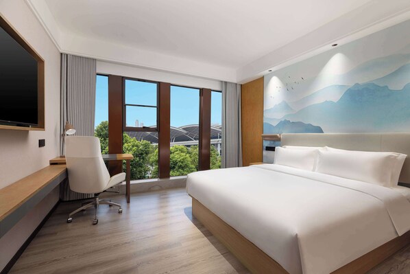 A hotel room with a large bed, a desk with a chair, and a view of greenery and a bridge through the window.