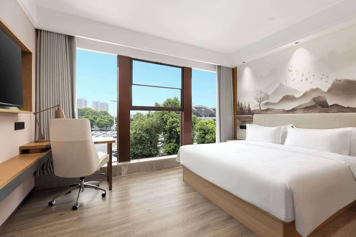 A modern hotel room with a large bed, a desk, and a view of a cityscape through large windows.