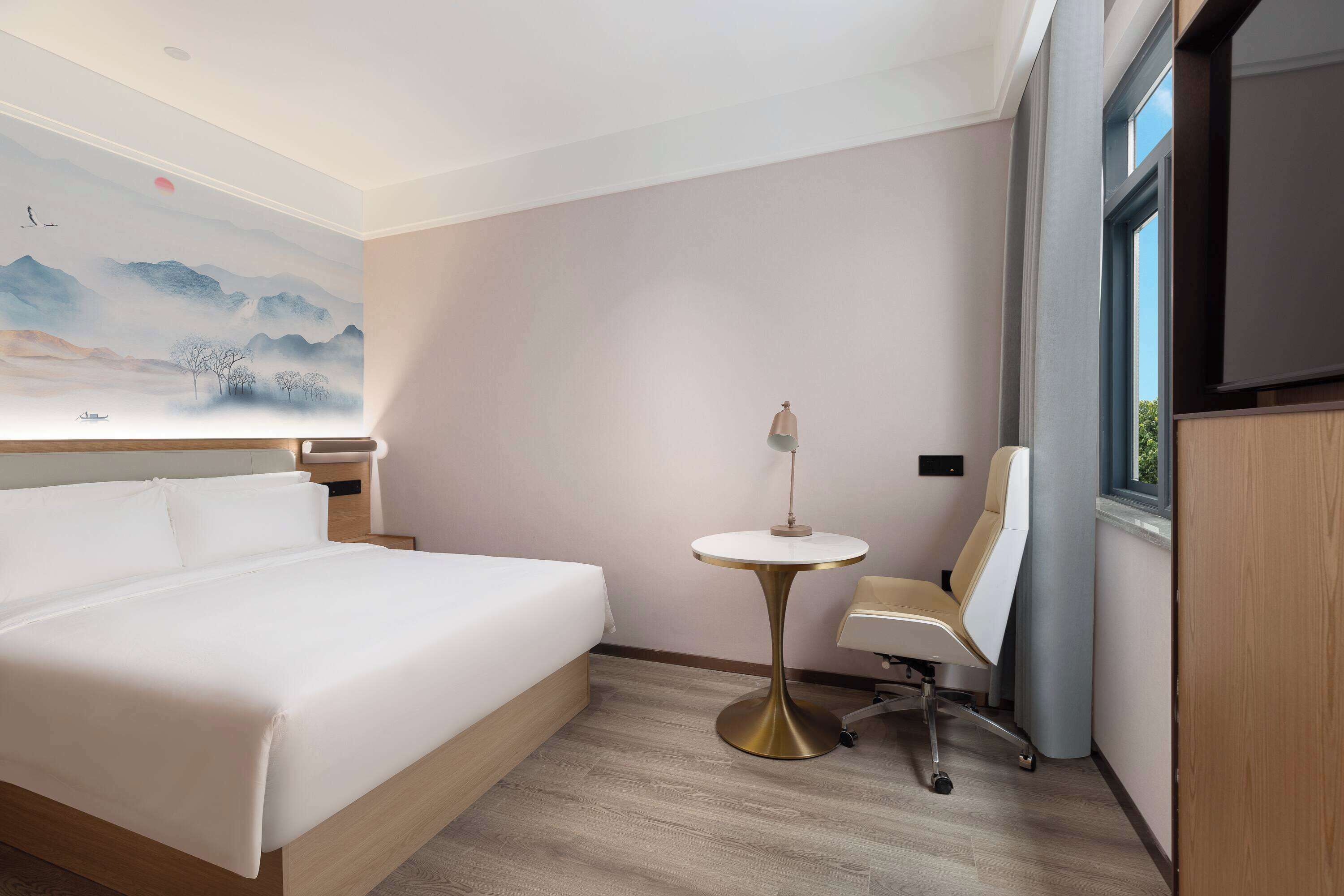 Una habitación de hotel con una cama grande, una mesita redonda pequeña, una silla y un cuadro en la pared.