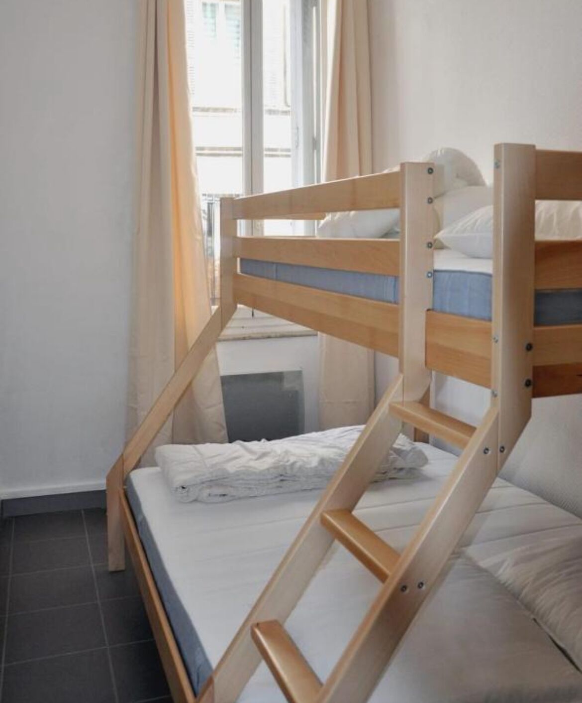 Una cama litera con escalera de madera, una mesita de noche pequeña y una ventana con cortinas.