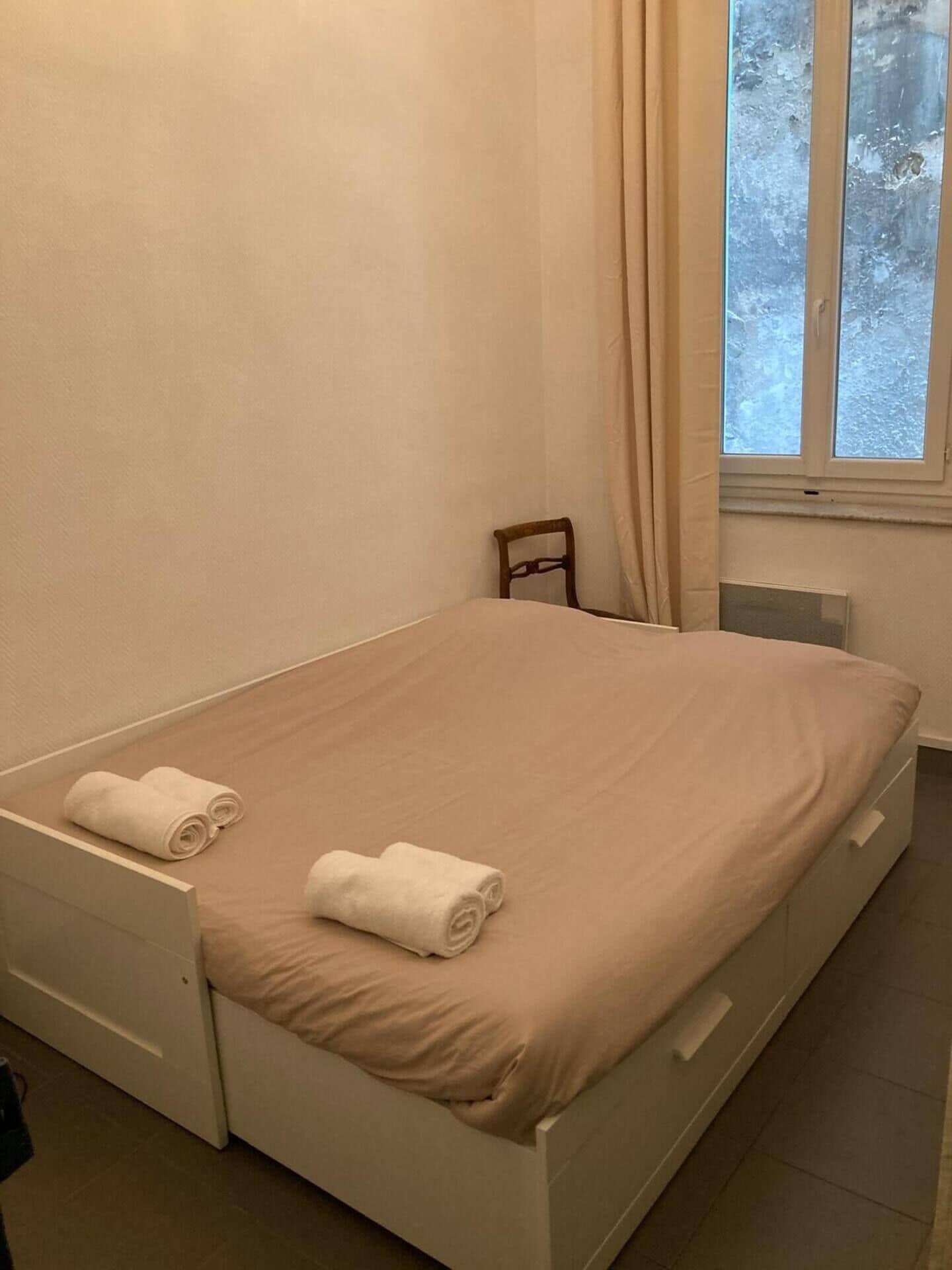 Une chambre avec un lit simple, une couvre-lit beige, deux serviettes enroulées, une chaise en bois et une fenêtre avec des rideaux.