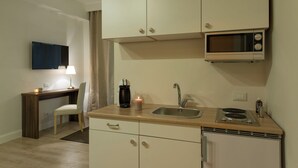 Eine kleine Küche mit Mikrowelle, Spüle und einem Herd. Ein Stuhl und ein Schreibtisch mit Lampe und Kerze sind ebenfalls vorhanden.