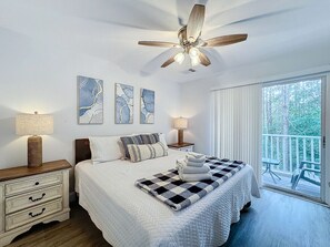 A bedroom with a bed, a ceiling fan, a nightstand, a lamp, and a view of the outdoors.