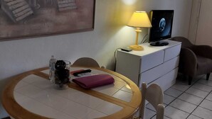 A room with a round table, chairs, a desk with a lamp, a TV, a painting of a beach scene, and a cabinet.