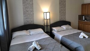 A hotel room with two beds, a nightstand, a lamp, a brick accent wall, and a kitchenette.