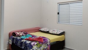 Une petite pièce avec un plafond en bois, un lit recouvert d’une courtepointe colorée, une fenêtre avec des stores et un luminaire.