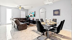 A modern living room with a sectional sofa, a dining table with chairs, a chandelier, and artwork on the walls.