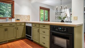 Una cocina con gabinetes verdes, lavavajillas, horno, fregadero y ventana con vista a árboles.