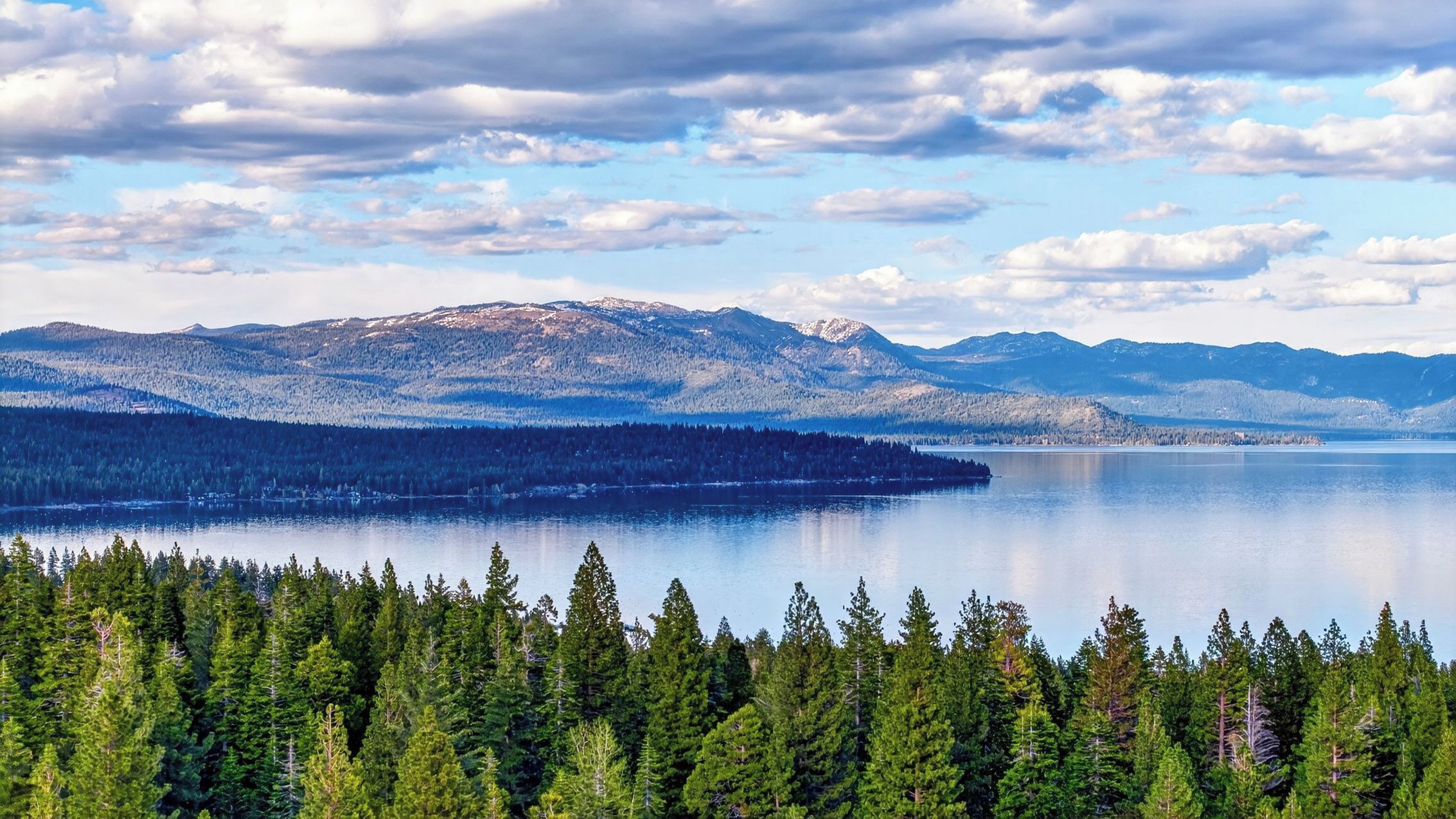 A lake surrounded by dense forests and snow-capped mountains under a partly cloudy sky.
