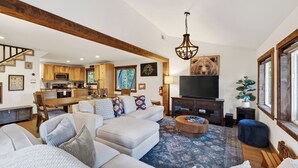 A spacious living room with a large sofa, a coffee table, and a television. There is a kitchen in the background with wooden cabinets and a dining area. - Washoe Way by Avantstay Luxury Tahoe Retreat A+ Location w/ Beach Access (Tahoe City)