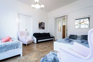 Un salon moderne avec un canapé blanc, un canapé noir, un fauteuil blanc et un fauteuil rose. Le sol est en bois et il y a un lustre.
