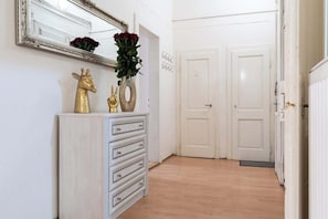 Um corredor com cômoda branca, espelho e um vaso de flores vermelhas. Há duas portas brancas e um radiador à direita.