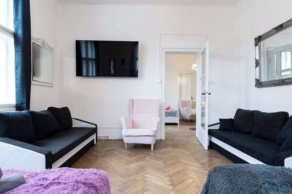 A modern living room with a herringbone floor, two black sofas, a pink armchair, a large flat-screen TV, and a mirror.