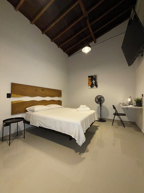 Quarto com cabeceira de madeira, cama com roupa de cama branca, escrivaninha com um laptop e uma planta, ventilador de chão e uma cadeira.