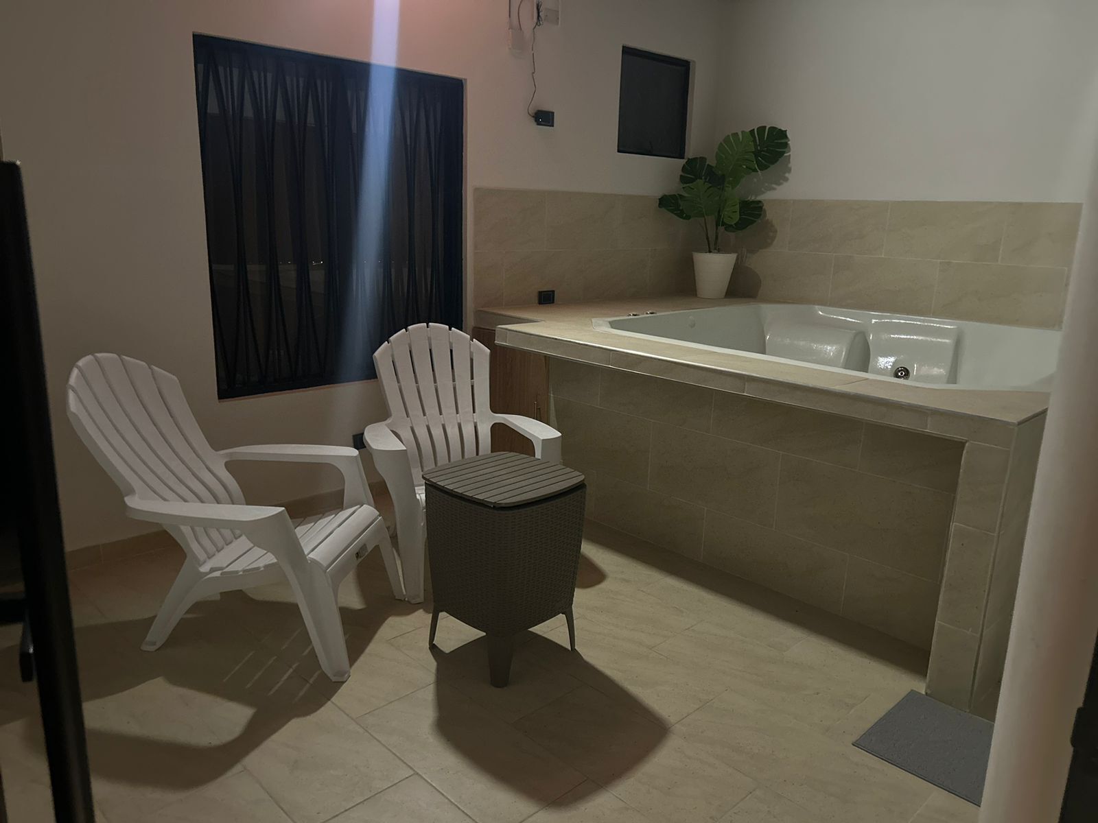 A small, modern bathroom with a bathtub, two white chairs, a small table, and a potted plant.