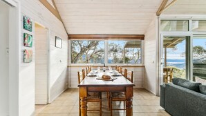 A dining area with a wooden table and chairs, large windows with a view of trees and hills, and a grey sofa. - Silvertop Apartment capturing the beautiful mountain and lake views (Crackenback)
