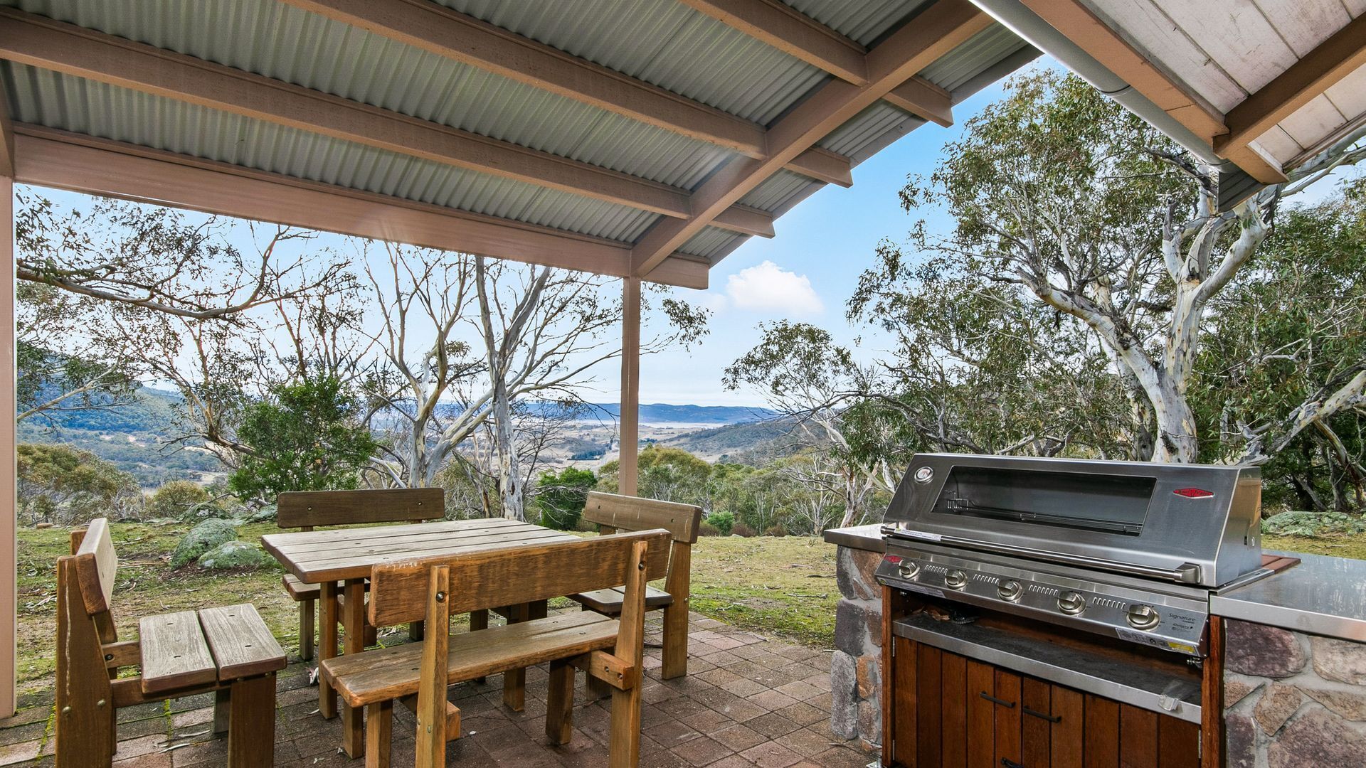 An outdoor patio with a wooden table and benches, a barbecue grill, and a view of the surrounding landscape.