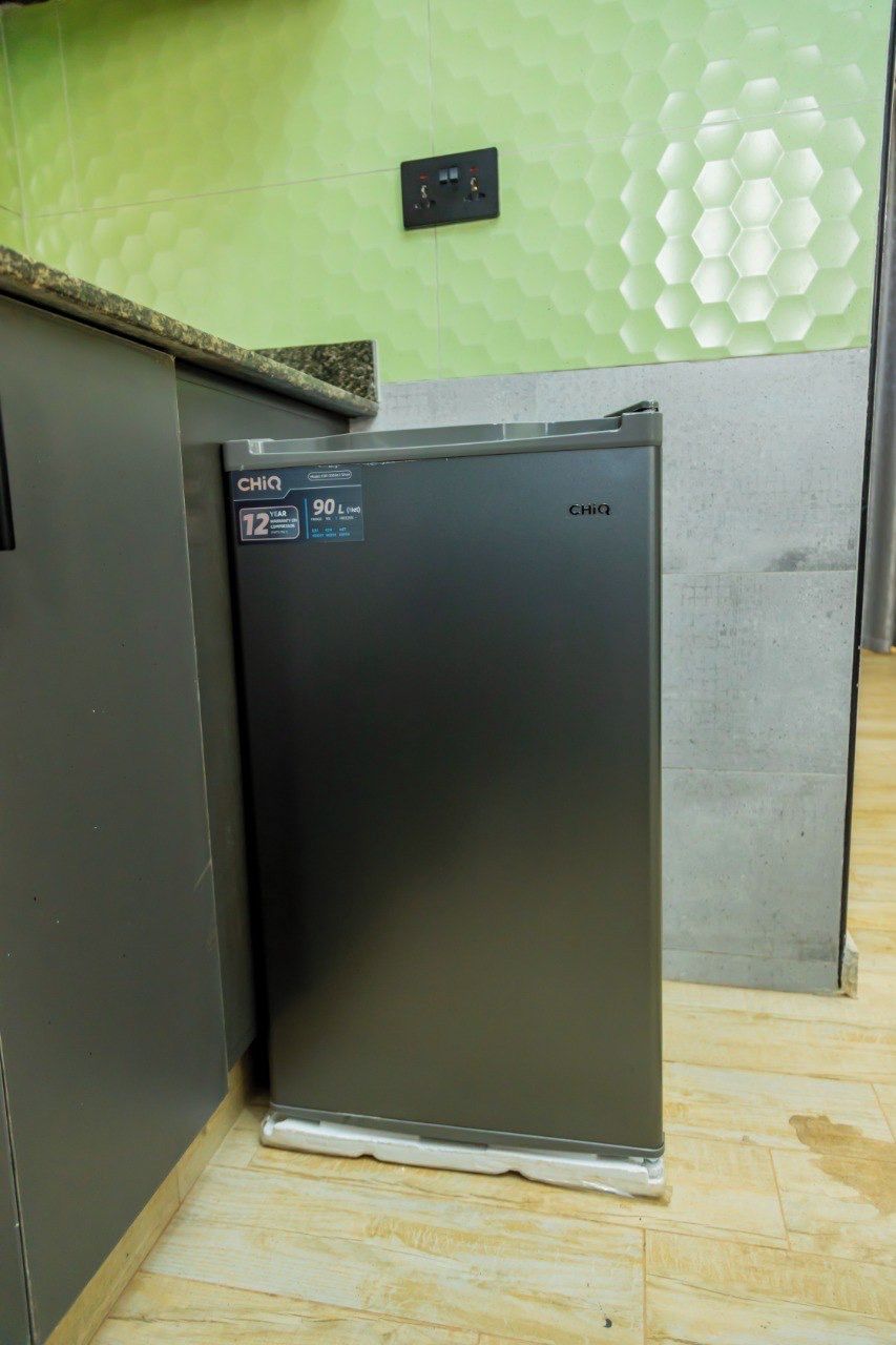 A compact refrigerator with a digital display, situated in a corner of a room with a green textured wall and a grey floor.